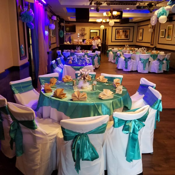 Restaurant dining room set for an event with teal tablecloths, white chair covers, and gold napkins.