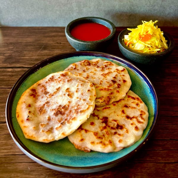 Three golden-brown pupusas on a teal plate, with red sauce and curtido in small bowls.