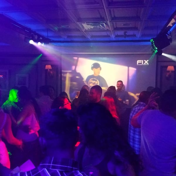 People dancing in a dark room with colorful lights and a DJ projected on a screen.