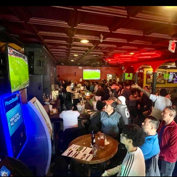 A lively crowd watches soccer on multiple screens in a dimly lit bar with red neon lights.