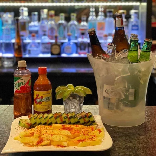 A plate of sliced cucumbers and mangoes with chili powder, hot sauce, and a bucket of beer.