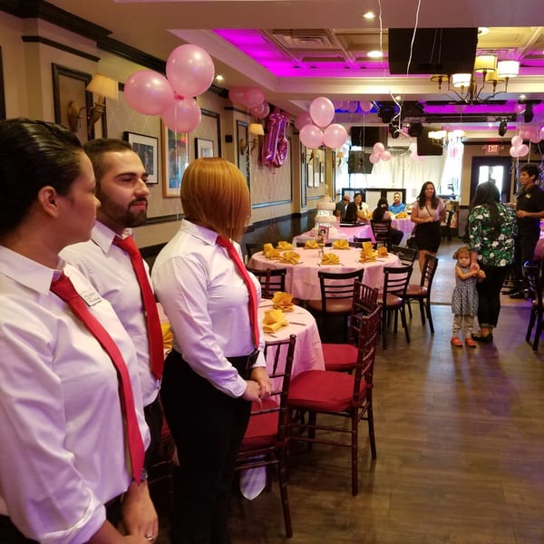 Restaurant staff in white shirts and red ties stand in a decorated dining room with pink balloons.