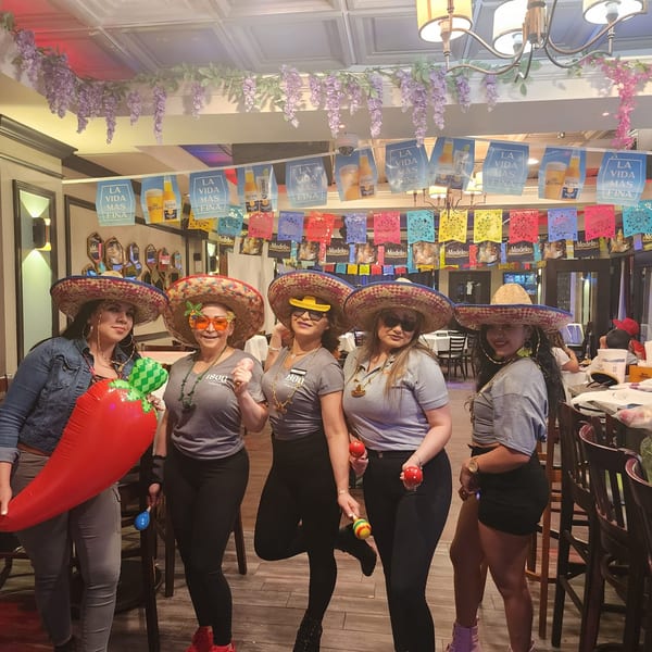 Five women in sombreros and festive attire pose with props in a decorated Salvadoran restaurant.