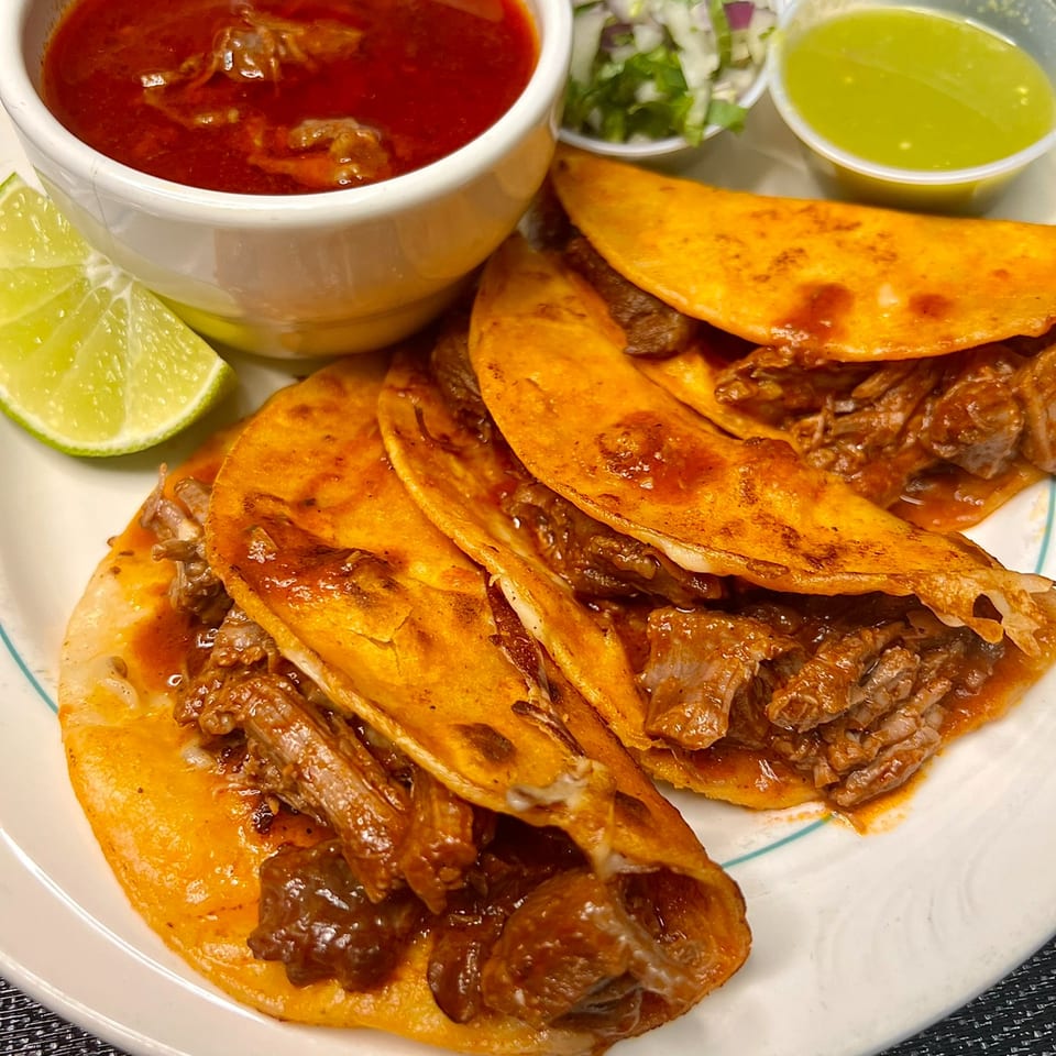Three birria tacos with a side of consommé, lime, and two sauces on a white plate.
